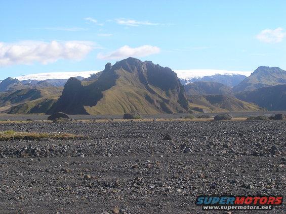 Mountain landscape with a rocky foreground and a glacier in the background. Supermotors.net logo in the lower right