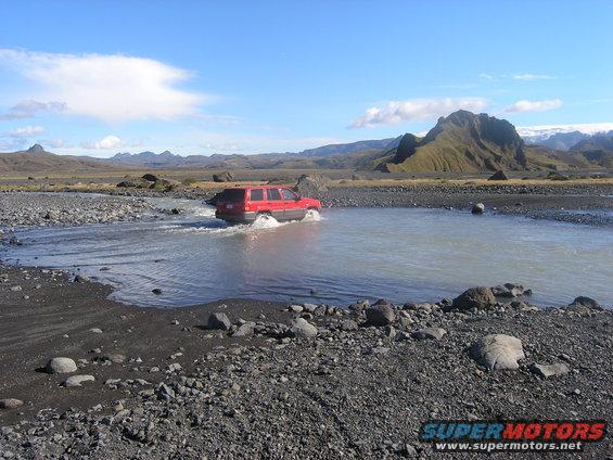 Red SUV driving through a shallow river in a rocky, barren landscape with mountains in the background