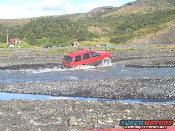 Red SUV crossing a rocky riverbed with hills in the background