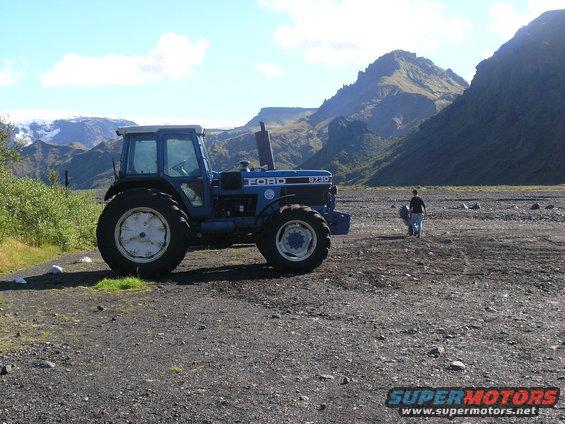 Blue Ford tractor with white rims parked on gravel, mountains in background