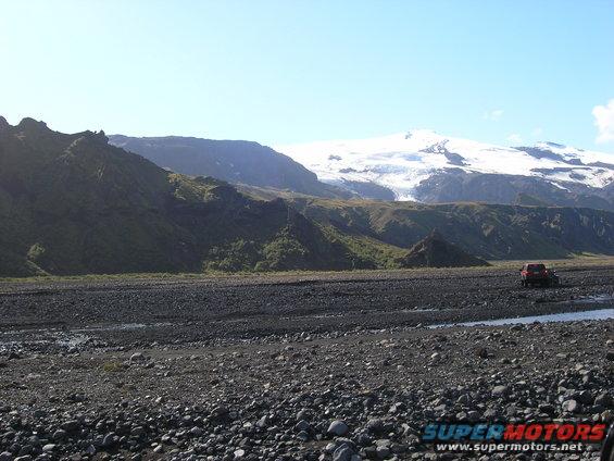Red SUV driving across a rocky, barren landscape with mountains and a glacier in the background
