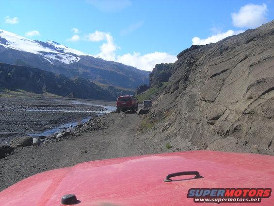 Two red SUVs drive on a dirt road through a rocky valley with a snow-capped mountain in the background