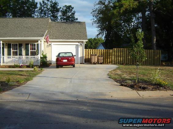 Red sedan parked in front of a garage. House has a porch with white railing and an American flag