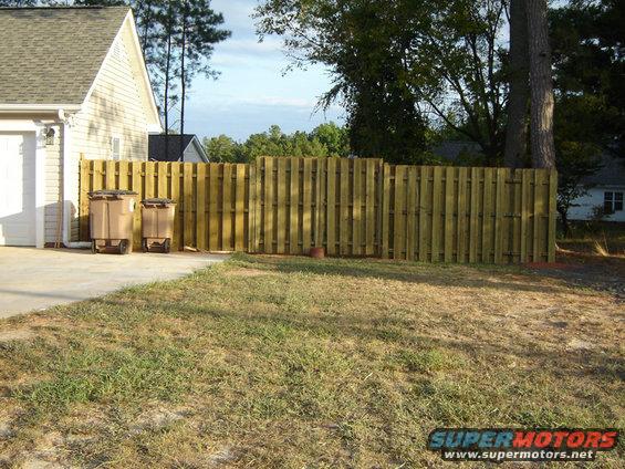 Two tan trash cans on a concrete driveway next to a wooden fence