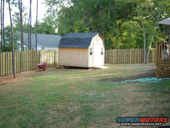 Backyard scene with a wooden shed, a red wheelbarrow, and a wooden fence