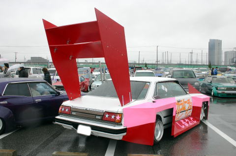 Modified white sedan with a huge red wing and pink/red body kit, parked in a lot with other cars