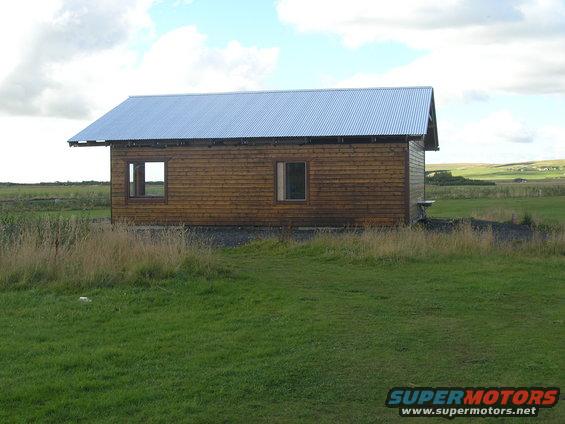 Wooden cabin with a metal roof in a grassy field under a cloudy sky. Supermotors.net logo in the corner