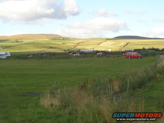 Red van and white RV parked in a field near cabins, with rolling hills in the background