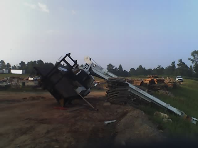 Overturned truck with crane in a dirt lot, with stacks of lumber and a white pickup truck in the background
