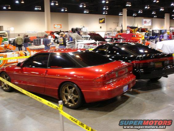 Red sports coupe with gold rims, rear 3/4 view, at an indoor car show