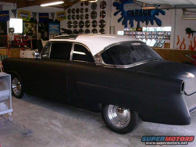 Black coupe with white roof, chrome trim, and custom rear end, parked in a garage with license plates on the wall