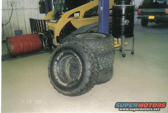 Four Yokohama Geolandar M/T tires stacked in front of a yellow CAT skid steer loader. A black Ford Bronco is visible in the background