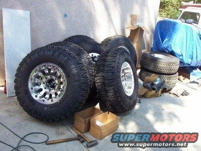 Four off-road tires with chrome rims piled on the ground next to a vehicle covered in a blue tarp
