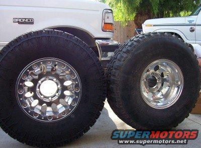 Two large off-road tires with chrome wheels are displayed in front of a white Ford Bronco and another white Ford truck