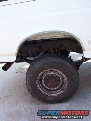 White SUV's rear wheel and leaf spring suspension, showing a rusty frame and exhaust pipe