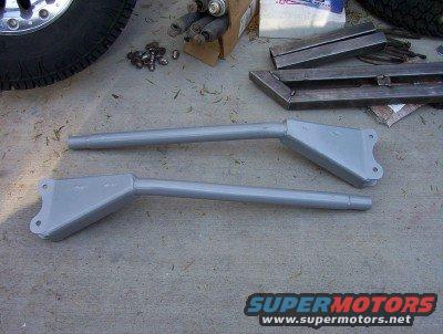 two gray metal suspension control arms on concrete, tire and metal parts in background