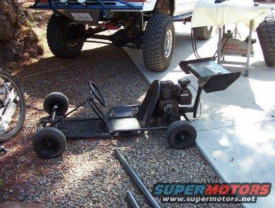Black go-kart with a small engine and a bicycle wheel nearby, parked on gravel next to a large truck