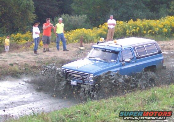 Blue SUV with roof lights driving through a mud pit, people watching