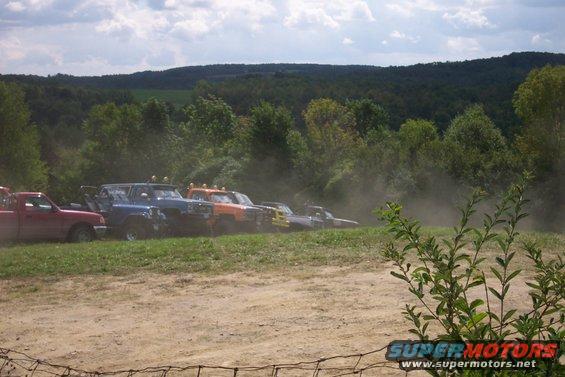Several off-road vehicles parked on a grassy incline, kicking up dust under a cloudy sky