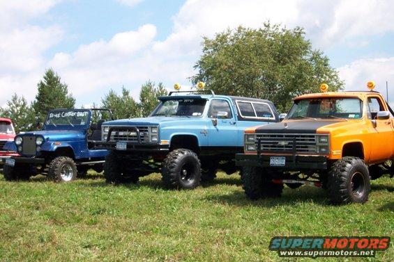 Orange and blue lifted trucks parked on grass, one with a black hood