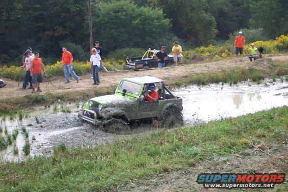 Green Jeep partially submerged in mud, with people watching and another truck in the background