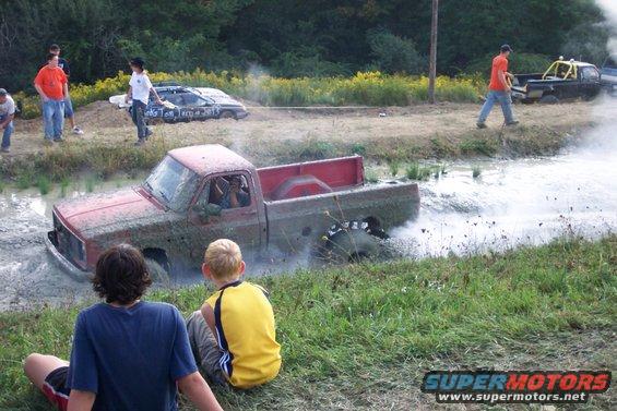 Red 1979 Ford F-150 stuck in mud pit with spectators watching
