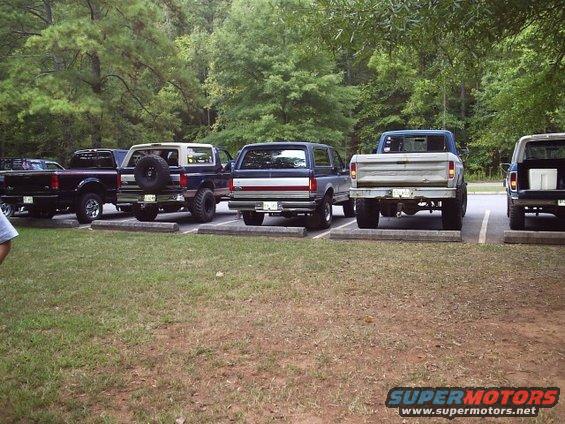 Several Ford trucks and a Bronco parked in a grassy lot with trees in the background. The Bronco appears to be from the late 1980s