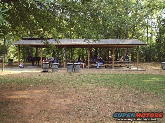 Park pavilion with picnic tables and people gathered under it, surrounded by trees
