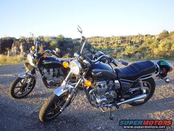 Two dark motorcycles parked on a gravel road, one with a green helmet on the back