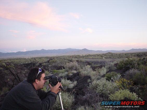 Man filming landscape with camera on tripod, mountains in background, sunset sky