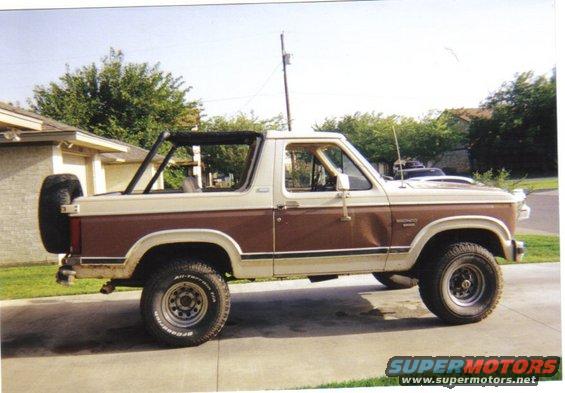 Side view of a two-tone brown and tan Ford Bronco with a spare tire on the back. It has a roll bar inside