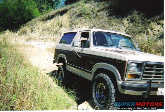 1980s Ford Bronco driving on a dirt path with dry grass and trees