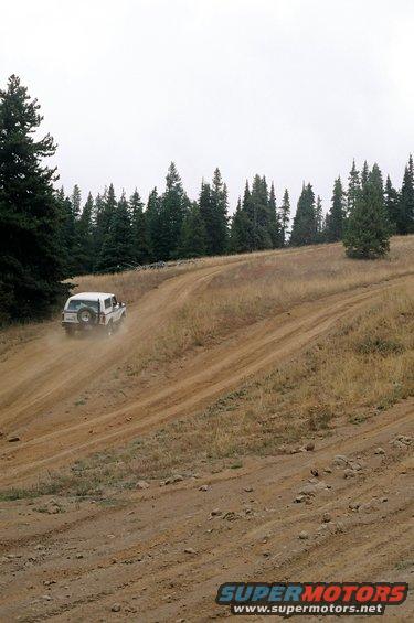 White 1979 Ford Bronco driving on a dirt road with trees in background