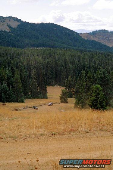 White SUV parked in a grassy field with a forest and mountains in the background