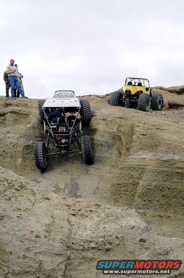 Two custom off-road vehicles navigate a steep, rocky terrain under an overcast sky