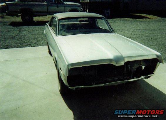 Silver coupe with missing grille and headlights, parked on concrete. A pickup truck is visible in the background