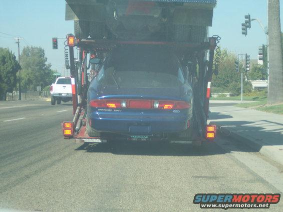Rear view of a blue 1995 Ford Probe on a car carrier, with a white truck visible ahead