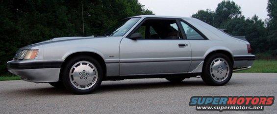 Silver 1986 Ford Mustang coupe parked on asphalt with trees in background