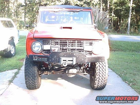Red 1974 Ford Bronco front view with winch and "Low 4x4 Country" sticker on fender. Another white SUV is parked to the left