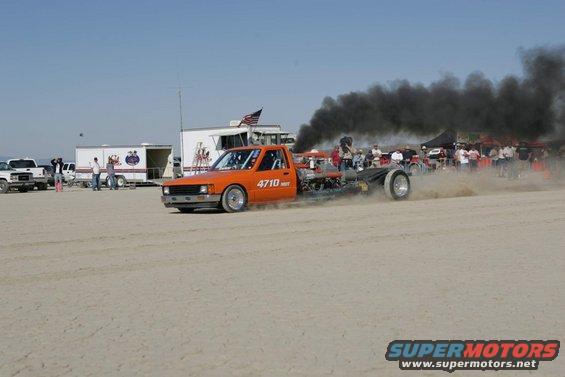 Orange pickup truck with custom chassis and large exhaust emitting black smoke, seen from the side