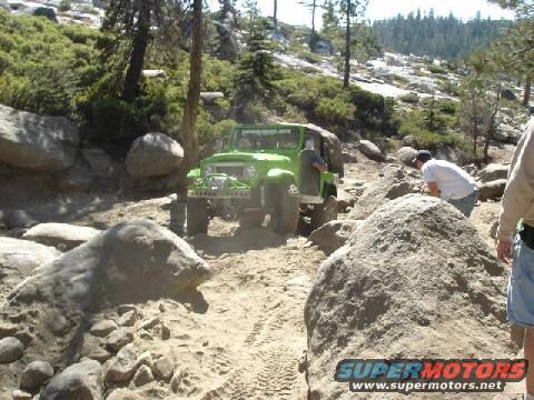 Green off-road vehicle navigating a rocky trail with people nearby