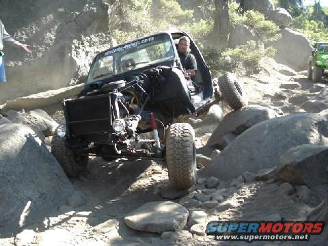 Off-road vehicle with open hood navigating rocks; green vehicle in background