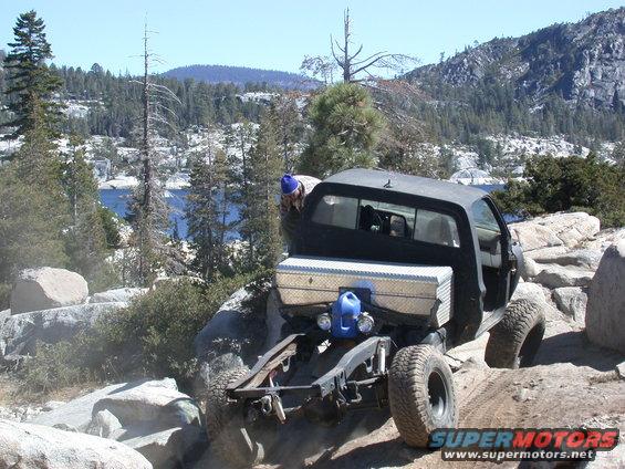 Pickup truck with a toolbox and gas can on the back, driving over rocks near a lake