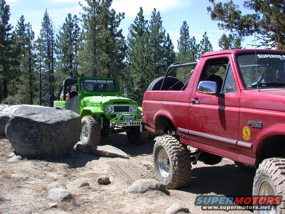 Red 1996 Ford Bronco and green off-road vehicle on a rocky trail with pine trees