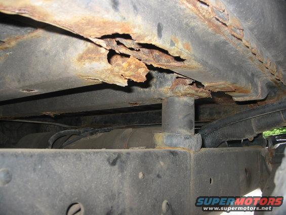 Heavily rusted frame rail and crossmember on a vehicle, showing significant corrosion and structural damage. A fuel tank is visible below