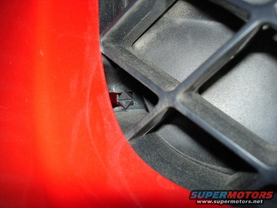 Close-up of a black plastic grille with a red panel on the left. A small black clip is visible in the grille opening. Supermotors.net logo in the corner
