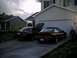 2004 Ford Mustang - Two sports cars, a red one and a black one with its trunk open, are parked in a driveway next to a house.