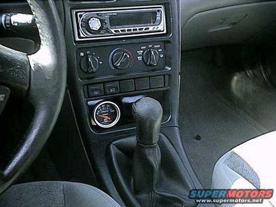 Car interior showing steering wheel, dashboard with radio, climate controls, gear shifter, and an aftermarket gauge