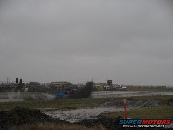 Blue pickup truck kicking up mud during a race, with other vehicles and spectators in the background under a cloudy sky