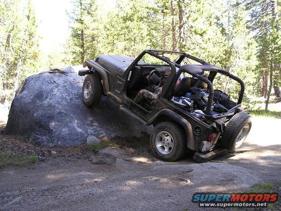 Jeep Wrangler TJ climbing over a large rock on a dirt trail. Rear-mounted spare tire is visible
